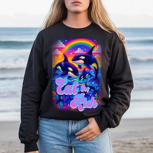 Puede incluir: Sudadera negra con un gráfico vibrante de orcas bajo un arcoíris. El diseño incluye el texto "Eat the Rich" en una fuente colorida y estilizada. Las orcas están representadas en un estilo de dibujos animados, con un fondo oceánico brillante y colorido.