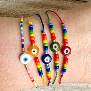 May include: Five colorful beaded bracelets with evil eye charms. The bracelets are made with small, colorful beads and feature a variety of evil eye charms in different colors, including blue, green, and yellow.