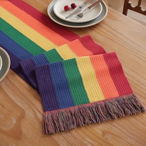 Könnte beinhalten: Ein Regenbogen-Tischläufer mit Fransen. Der Läufer hat Streifen in Rot, Orange, Gelb, Grün, Blau und Lila. Der Läufer besteht aus einem strukturierten Stoff und wird auf einem Holztisch präsentiert, mit Tellern und Besteck im Hintergrund.