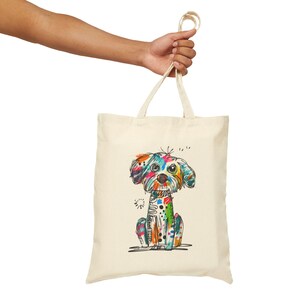 Könnte beinhalten: Cremefarbene Tragetasche mit einer farbenfrohen, skurrilen Illustration eines Hundes. Der Hund ist mit einer Vielzahl von leuchtenden Farben dargestellt, darunter Blau, Orange, Grün und Rosa. Die Tasche hat zwei Griffe und wird von einer Person gehalten.