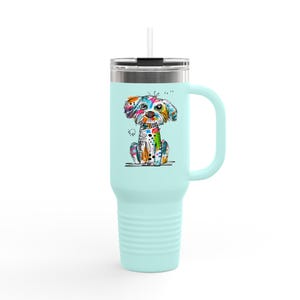 May include: A pale blue travel mug with a handle and a straw. The mug showcases a vibrant illustration of a dog with a playful design. The dog is rendered in a spectrum of colours, including blue, green, yellow, and orange.