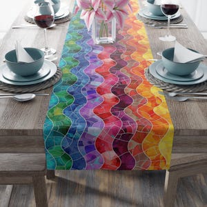 May include: A colorful table runner with a mosaic-like design in a spectrum of colors, including green, blue, purple, red, orange, and yellow. The wavy pattern adds visual interest to the dining table setting. The table is set with plates, bowls, and wine glasses.