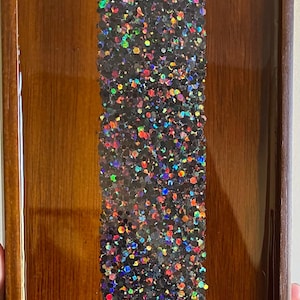 May include: A wooden tray with a vertical strip of black resin embedded with iridescent glitter. The glitter is a mix of red, orange, yellow, green, blue, and purple.