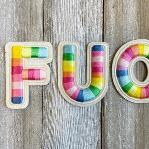 May include: Three embroidered felt patches in the shape of the letters F, U, and O. The patches are made of a white felt base with colorful embroidery in shades of pink, yellow, blue, and green. The letters are arranged in a row, spelling out the word "FUO".