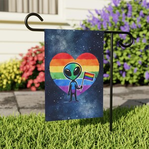 May include: A garden flag with a cartoon alien holding a rainbow flag, set against a starry night sky. The alien is inside a rainbow heart. The flag is displayed on a black metal stand, set in a grassy garden.