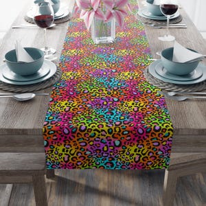 Puede incluir: Un vibrante camino de mesa con un diseño de estampado de leopardo colorido. El patrón presenta una mezcla de colores brillantes que incluyen manchas rosas, amarillas, naranjas, verdes y azules sobre un fondo negro. El camino de mesa se muestra sobre una mesa de madera, preparada para una comida.