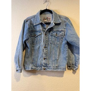 May include: Light blue denim jacket with a classic collar, button-down front, and two chest pockets. The jacket has long sleeves with buttoned cuffs and a small patch on the upper left chest. The jacket is hanging on a black hanger.