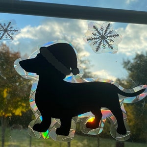 May include: A black silhouette of a dachshund wearing a Santa hat, with a sparkly iridescent background. The image is a window sticker with two snowflakes.