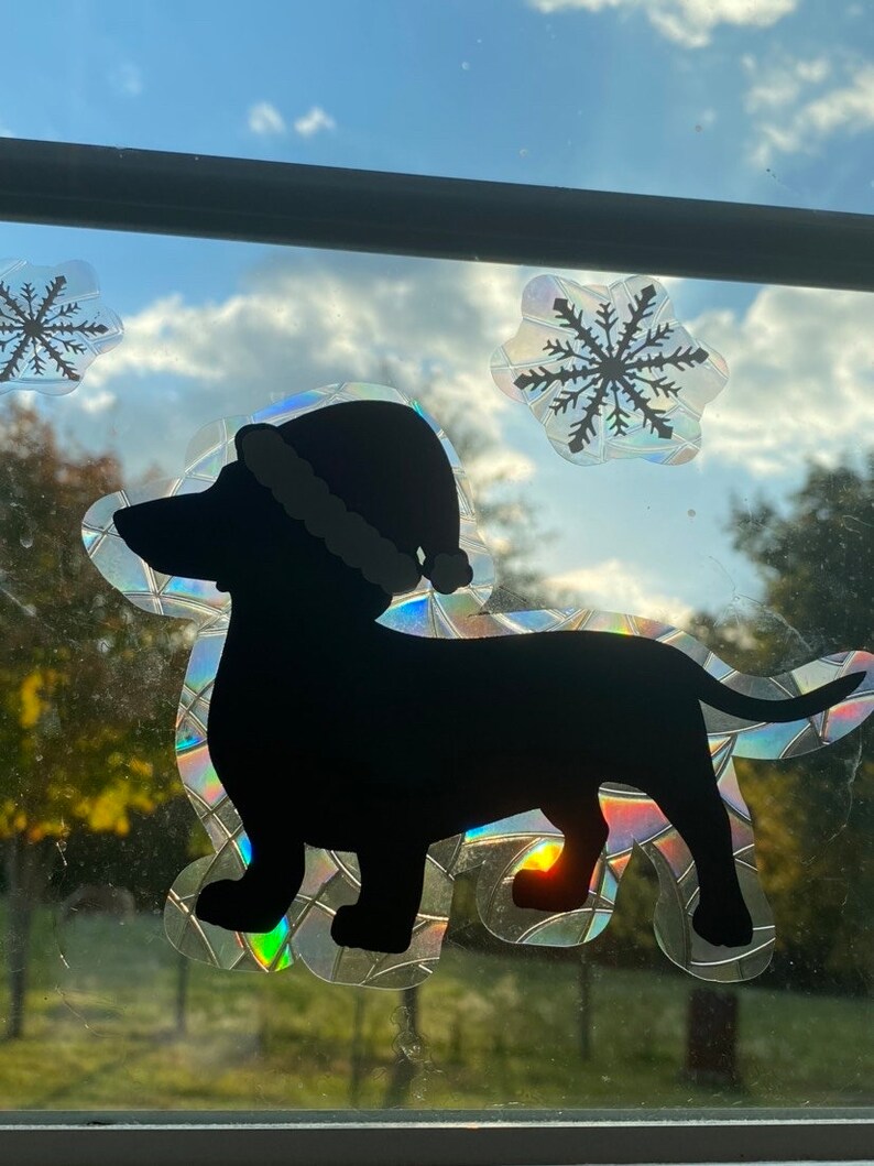 May include: A black silhouette of a dachshund wearing a Santa hat, with a sparkly iridescent background. The image is a window sticker with two snowflakes.