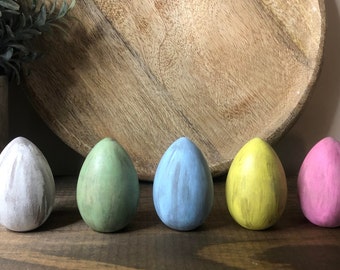 May include: Five wooden eggs painted in different colors. The eggs are arranged in a row from left to right: white, green, blue, yellow, and pink.