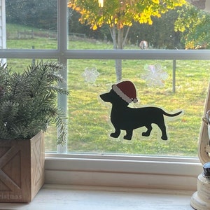 May include: A black silhouette of a dachshund wearing a red Santa hat is on a window with a frosted snowflake design. The window is behind a potted evergreen plant and a white lantern.