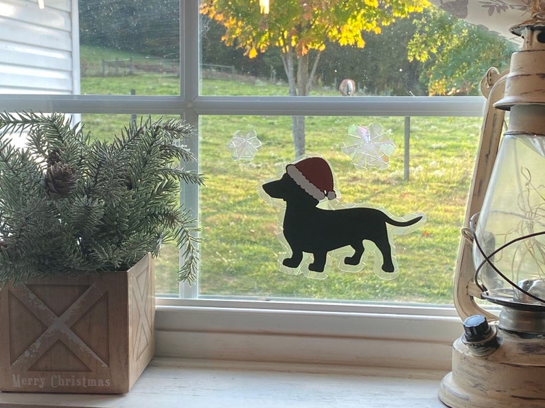 May include: A black silhouette of a dachshund wearing a red Santa hat is on a window with a frosted snowflake design. The window is behind a potted evergreen plant and a white lantern.