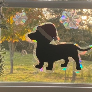 May include: A black silhouette of a dachshund wearing a red Santa hat, with two iridescent snowflake stickers on a window.
