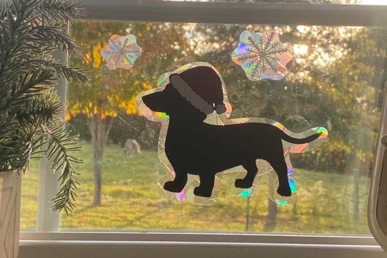 May include: A black silhouette of a dachshund wearing a red Santa hat, with two iridescent snowflake stickers on a window.
