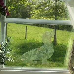 May include: A stained glass window cling of a dachshund dog silhouette, with a green, blue, and white mosaic pattern, against a backdrop of a green grassy field.