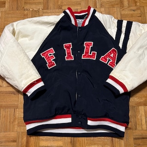 May include: A navy blue and cream-colored Fila letterman jacket. The jacket features the word "FILA" in red block letters across the chest. The sleeves are cream-colored, and the jacket has red and white striped trim at the cuffs and waist.