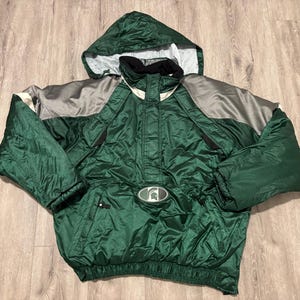 May include: Green and grey hooded anorak jacket with a front pocket and a logo. The jacket has a black collar and a drawstring at the bottom. The anorak is made of a shiny, water-resistant material.