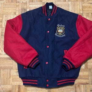 May include: A navy blue and red varsity jacket with a red and white striped collar and cuffs. The jacket has a large embroidered crest on the left side with the text "Oxford University".