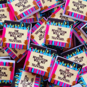 May include: A collection of square magnets with the text "THE REVOLUTION WILL NOT BE TELEVISED." Each magnet features a tan background with black, pink, and blue accents and a crowd silhouette.