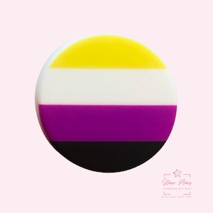 May include: A round, non-binary pride flag button with a yellow top stripe, white middle stripe, purple stripe, and black bottom stripe. The button has a glossy finish and is set against a light pink background. The button is a planner and gift shop item.