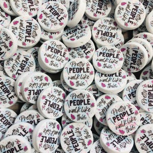 May include: A collection of white, round beads with the text "Pretty People Walk Like This" in black and pink. The beads have a small hole for stringing and a pink floral design. The beads are scattered.