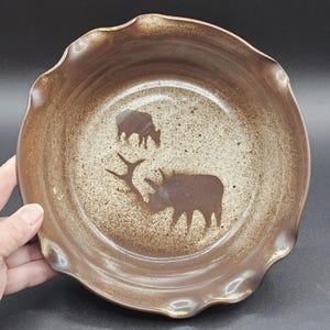 May include: A brown ceramic bowl with a ruffled edge. The interior features a speckled beige background with two dark brown elk silhouettes. The bowl is held by a hand.