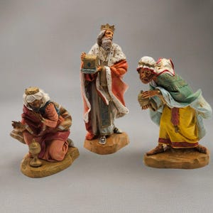 May include: Three detailed figurines depicting the biblical Magi. One kneels, another stands holding a box, and the third leans forward with a vessel. The figures are dressed in robes of red, gold, green, and blue, with intricate detailing and crowns. The figurines are on wooden bases.