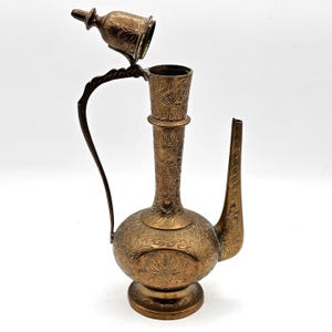 May include: An antique brass pitcher with intricate floral engravings. The pitcher features a long spout, a curved handle, and a hinged lid. The design includes a bulbous base and a tall, slender neck. The lid is open, revealing the interior.