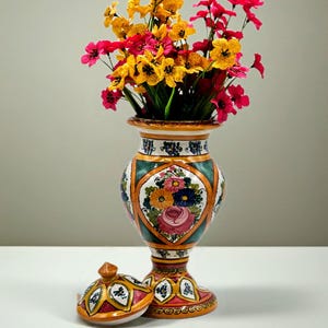 May include: A hand-painted ceramic urn-style vase with a lid, featuring a colourful floral motif in pink, yellow, and blue against a green and orange geometric background. The vase is filled with a bouquet of bright yellow and pink artificial flowers.