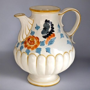 May include: Cream-colored ceramic pitcher with a floral design. The pitcher features a bulbous body with a scalloped base and a spout. The floral design includes orange roses, blue leaves, and black and orange accents. The handle and trim are accented with gold.