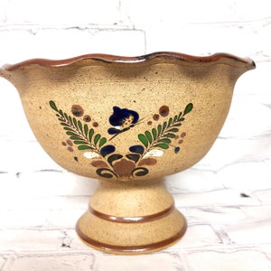 May include: A brown ceramic bowl with a scalloped rim and a pedestal base. The bowl is decorated with hand-painted green, blue, and white floral designs.