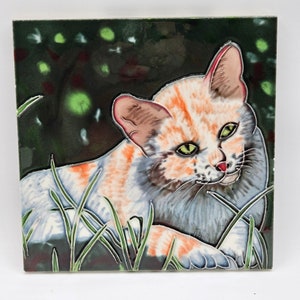 May include: A ceramic tile featuring a hand-painted illustration of a white and orange cat with green eyes, lying in tall green grass. The cat's fur is detailed with orange and white markings. The background is a dark green with a blurred pattern of green and white dots.