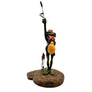 May include: A whimsical frog figurine with a green body, yellow belly, and a red scarf. The frog stands on a stone base, holding a cattail stalk. The figurine is a decorative item.