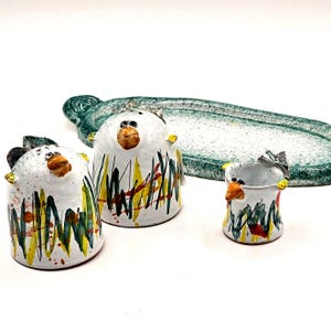 May include: A ceramic salt and pepper set and a small dish. The salt and pepper shakers are white with abstract yellow, green, and red designs. The dish is green with a speckled pattern. The set has a whimsical, artistic style.