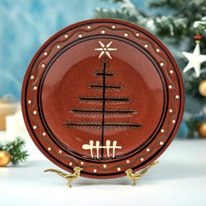 May include: A round, red ceramic plate with a hand-painted Christmas tree design. The plate features a black border with white dots and a decorative outer edge. The plate is displayed on a gold-colored bird-shaped stand.