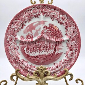 May include: A divided plate with a red and white floral and landscape design. The plate features a central scene of a horse-drawn carriage in front of a building, surrounded by a floral border. The plate is displayed on a decorative gold stand.