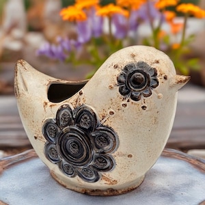 May include: A cream-colored ceramic bird-shaped planter featuring dark brown, embossed floral patterns on the side and eye area. The piece has a smooth, glazed finish with subtle speckling and a hollow opening at the top for small plants.
