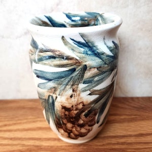 May include: A white ceramic vase with a brown and blue pine cone design. The vase is hand-painted with a stylized pine cone and branches.