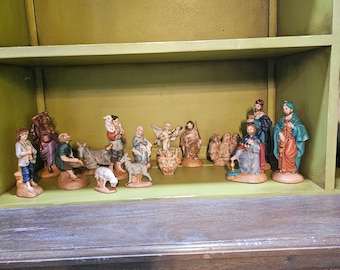 May include: A collection of hand-painted ceramic nativity figurines including figures of people, an angel, and various animals. The set features detailed clothing in muted tones, arranged on a shelf, depicting a traditional scene with multiple characters.