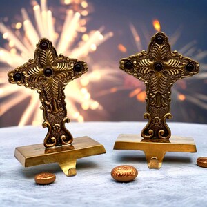 May include: Two decorative, gold-coloured cross-shaped stocking holders with intricate detailing and dark red accents. Each holder has a rectangular base with a hook. Small, gold-coloured, oval-shaped decorative pieces are scattered around the base.