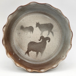 May include: A ceramic pie plate with a wavy rim and a speckled, neutral color. The plate features three dark brown silhouettes of sheep. The rim has a darker brown color, adding contrast to the design.