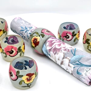 May include: Set of six gray napkin rings with hand-painted floral designs featuring pansies in shades of pink, yellow, and purple.