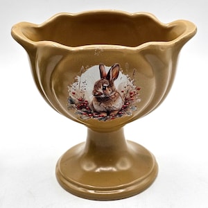 May include: A tan-colored ceramic pedestal bowl with a scalloped rim. The bowl features a detailed illustration of a brown and white rabbit surrounded by red berries and greenery. The bowl has a wide, rounded base.