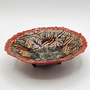 May include: A handmade ceramic bowl with a textured surface and a unique design. The bowl features a red rim and three small feet. The interior has a mix of brown, black, and blue colors, creating a striking contrast.