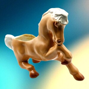May include: A ceramic horse planter in a light brown color with a white mane and tail. The horse is in a rearing pose with its front legs raised. The planter has a hollowed-out back for planting.
