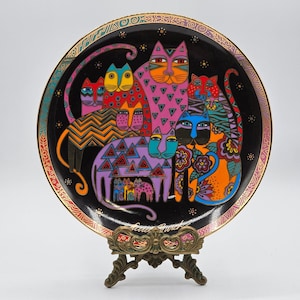 May include: A black ceramic plate featuring a colourful design of cats. The cats are all different colours and have various patterns on their fur. The plate is edged with a gold trim and comes with a gold stand.