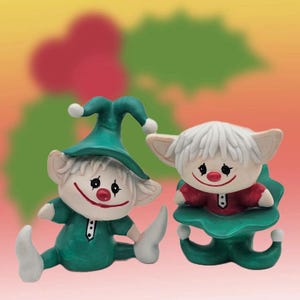 May include: Two ceramic elf figurines with white hair, red noses, and painted facial features. One elf wears a green pointed hat and outfit with white boots, while the other elf sits inside a green leaf-shaped base wearing a red shirt.