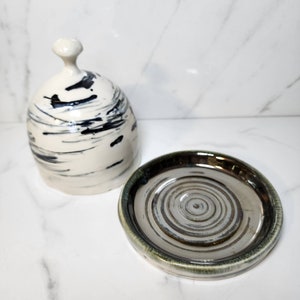 May include: A white ceramic butter dish with a black and white abstract design. The lid has a knob on top. The dish has a matching base with a metallic glaze.