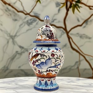 Peut inclure: Un pot en céramique avec couvercle amovible, orné d'un motif peint à la main représentant un oiseau bleu, des raisins violets et des vignes vertes sur fond blanc. Le récipient a un corps bombé, une base effilée et un bouton pointu sur le couvercle.