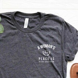 K&ouml;nnte beinhalten: Ein dunkelgraues T-Shirt mit dem Text "A Woman's Place is in the Constitution" und einer Grafik von einer Waage. Das Shirt wird mit einer blauen Jeans und braunen Sandalen kombiniert.